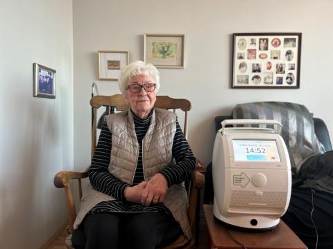 An older woman sits in a chair with a medication dispenser device beside her
