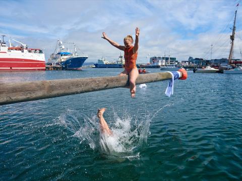 A young woman sits on a pole over the water and has successfully pushed another person into the sea. Nice weather, Reykjavík harbor, photo taken on Fishermen's Day