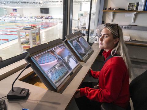 Lifeguard Dagbjört Ýr Gísladóttir monitors screens in the guard room at Laugardalslaug Pool. She's wearing a red sweater, with a window overlooking the indoor pool on the left side of the image.