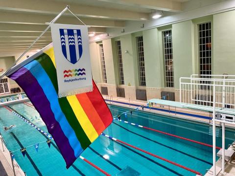The Rainbow Flag and the Reykjavík flag hang inside the Sundhöllin swimming pool, with the diving board and pool visible in the background