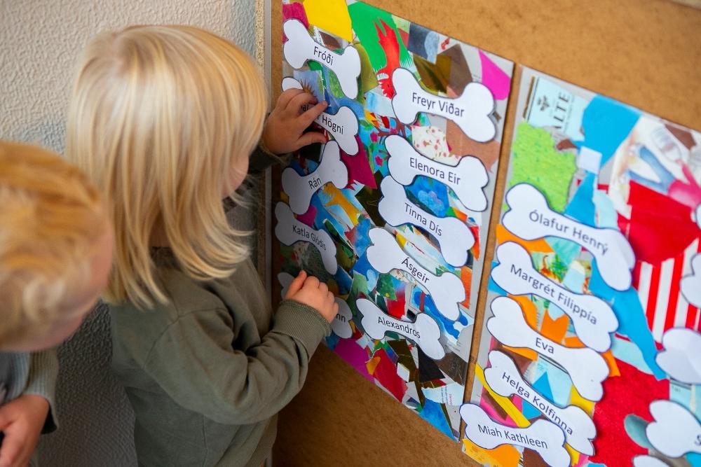 Fair-haired children looking at a poster on the wall where children's names are printed on white labels.