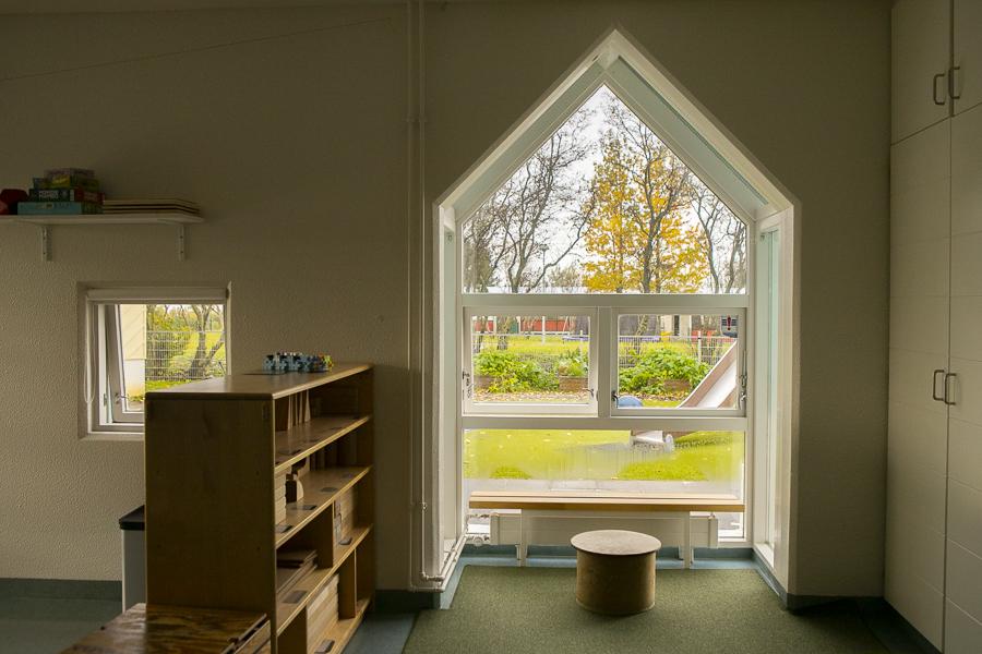 Looking out a window at Sólborg preschool. Inside is a shelf and a small chair; outside, a slide and other playground equipment.