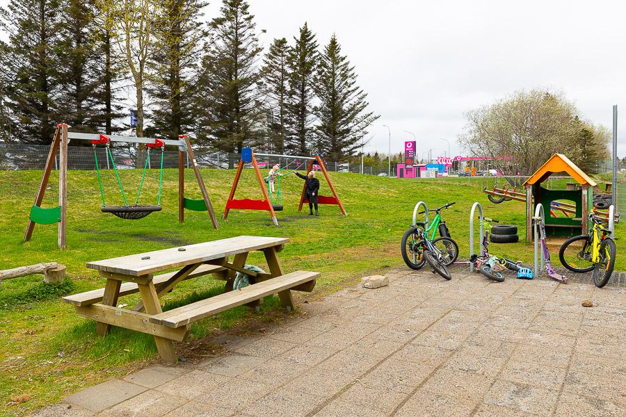 Outdoor play area with swings on a grassy slope, a table with benches, bicycles in a bike rack, and a small house. 