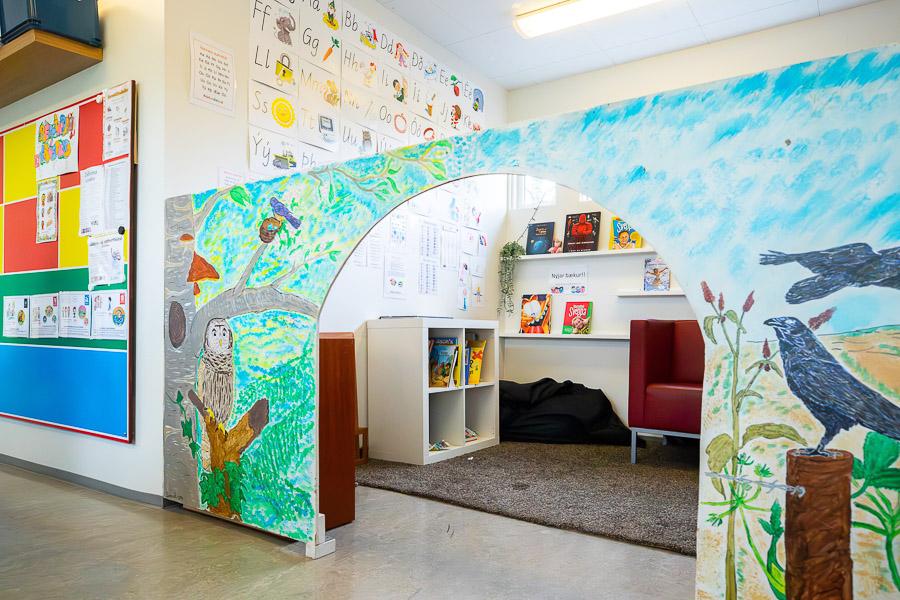 Seen through a painted partition wall decorated with pictures of trees and animals. A sofa and shelves are visible beyond the wall.