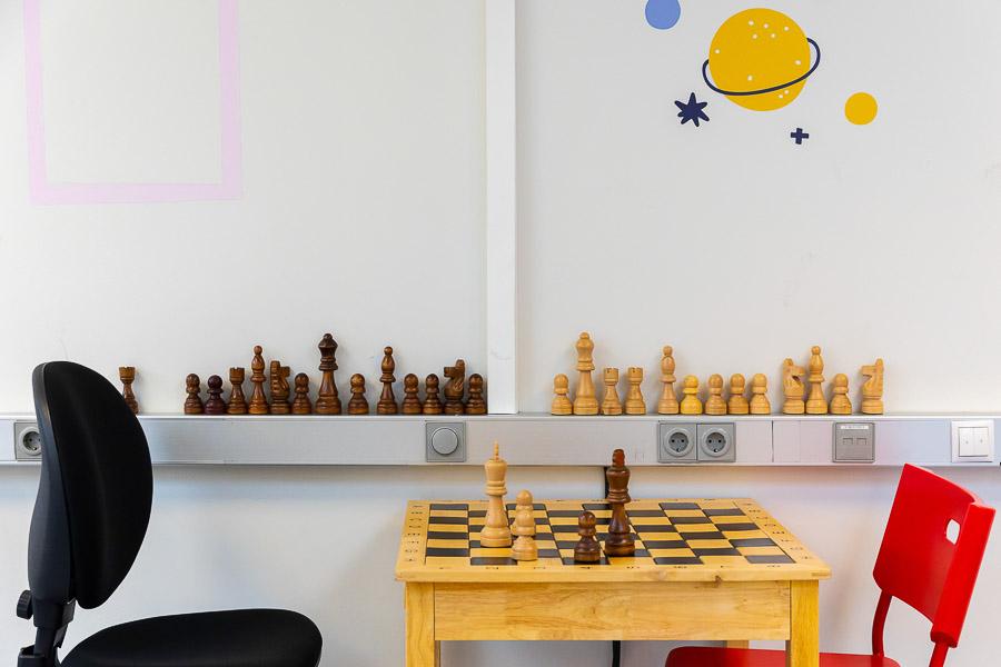 Chess table and two chairs. Chess pieces arranged on a shelf behind the table and planetary decorations on the wall.