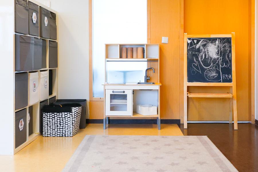 A toy kitchen and chalkboard in front of an orange wall. Shelves to the side.