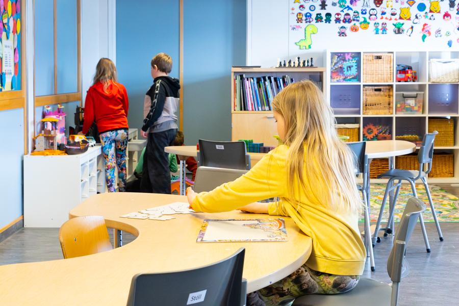 Child sits at a table working on a puzzle. Two children stand by a shelf playing with a dollhouse. Shelves with toys in the background.