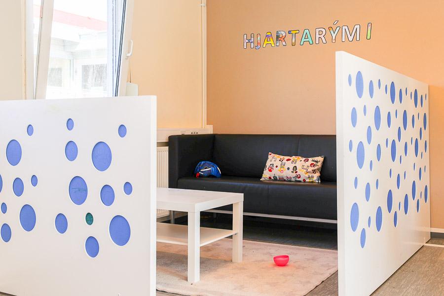 Light brown wall with the word Hjartarými (Heart Space). Black sofa, small table and rug. Low walls around the area with circular patterns. 