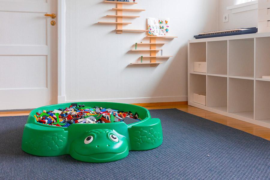 Large plastic turtle on the floor filled with Lego blocks. Shelves to the side and a gray rug on the floor. 