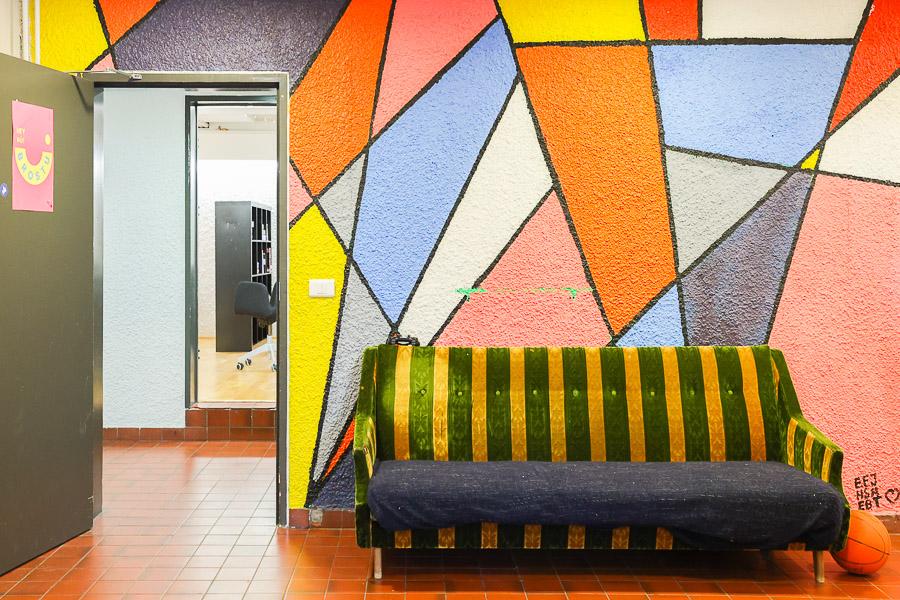 Wall painted with colorful patterns. Striped sofa in front with a door leading to the hallway on the side. 