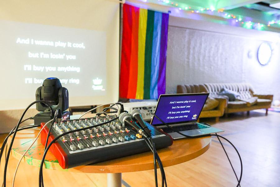 Computer, sound mixing board and microphones on a table in the foreground. Karaoke lyrics displayed on screen. LGBT+ flag hanging to the side next to the sofa. 