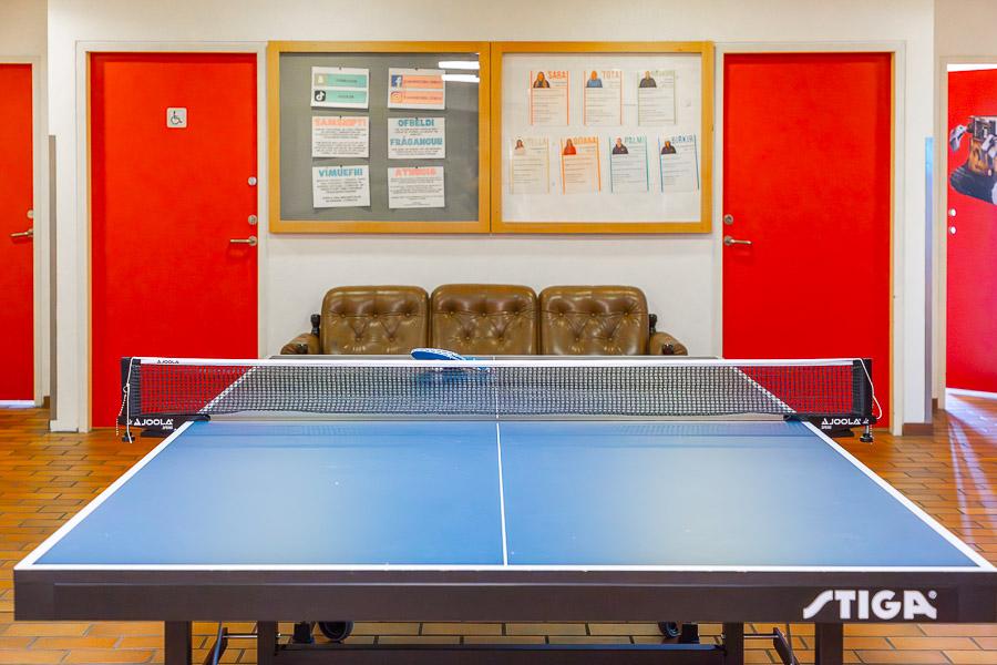 Room with a ping pong table, couch and notices on the wall. Red doors in the background. 