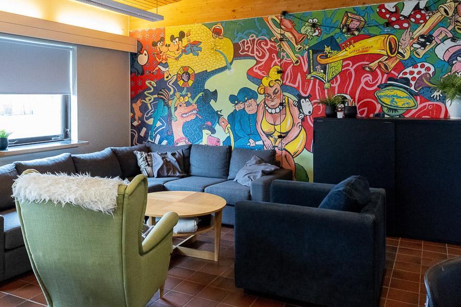 A large mural in the background. A corner couch, a coffee table, and chairs. A black shelf. 