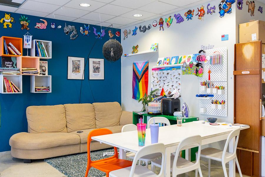 A sofa, table and chairs. A blue wall with shelves and pictures. An LGBT+ flag and Perler bead artwork on the wall.