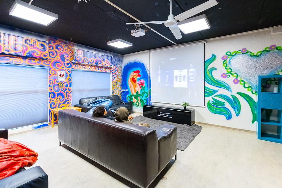 Young people sit on a sofa watching a screen. Surrounding walls with graffiti.
