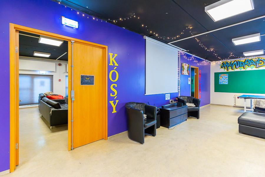 Purple wall with "Cozy" written on it. Black sofas and chairs. String lights hanging from the ceiling.