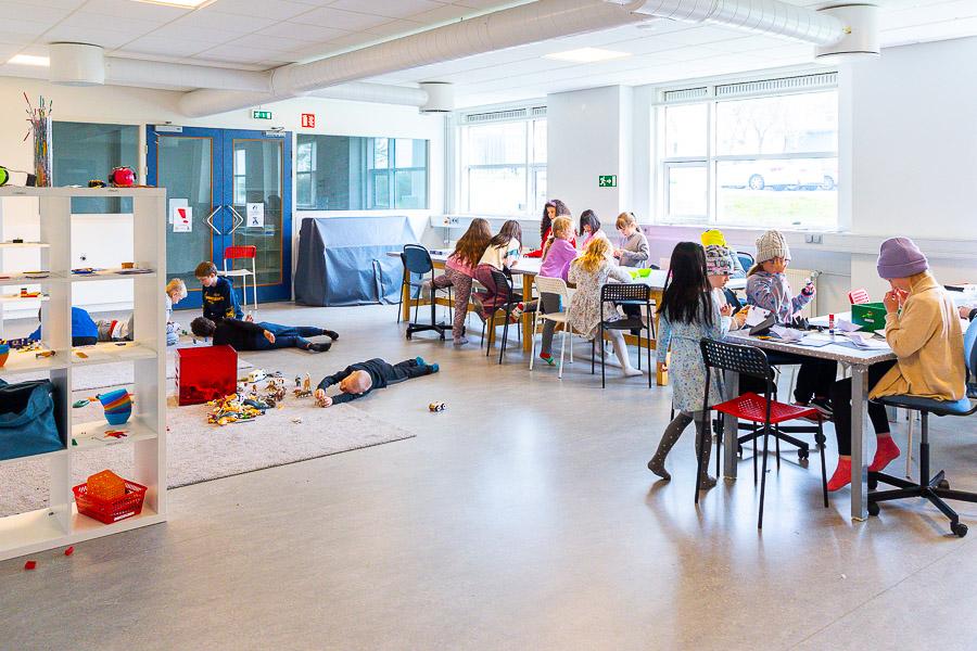 Children sit at two tables crafting. Other children are on the floor playing with toy animals and more. A white shelf is off to the side.