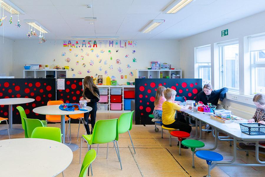 A hall with tables and chairs. Children sit on colorful stools, crafting. Room dividers with a circular pattern are in the background. "Children's Rights" is written on the wall.