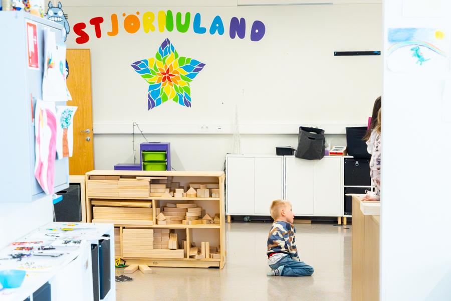 A child sits on the floor in a playroom. Shelves with large wooden blocks are on the side. On the wall is a large star and colorful letters forming the word "Stjörnuland." 