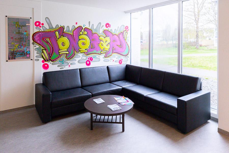 Black corner couch under the window and round coffee table. Tónabær has been painted on the wall. 