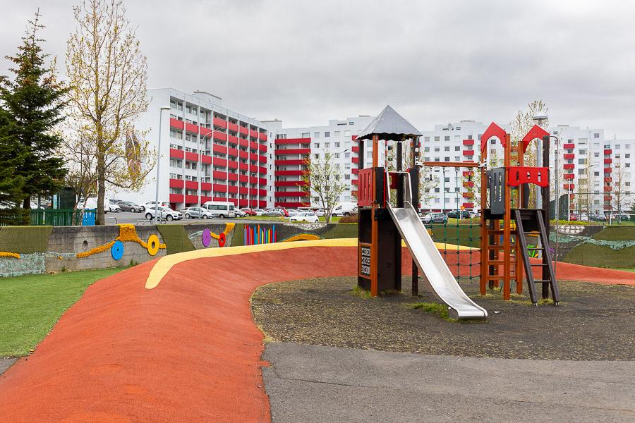 A playground featuring a climbing structure and slide. Apartment buildings visible in the background.