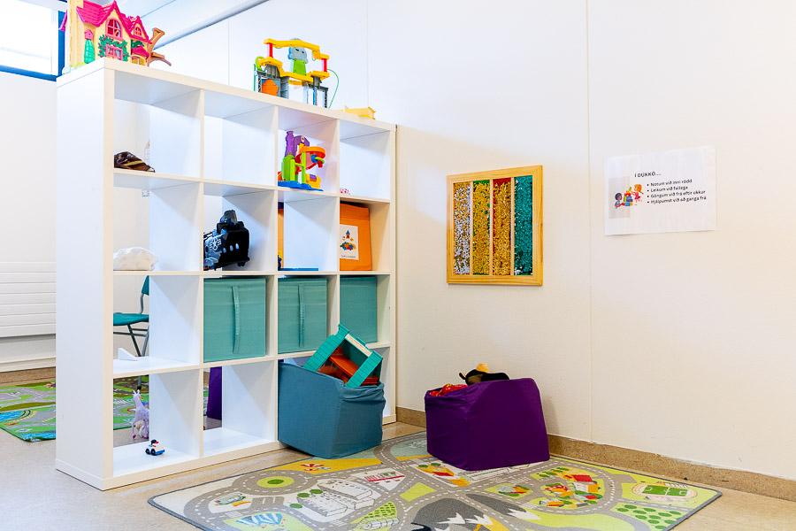 A white shelf filled with toys and green storage bins. A play mat featuring a road pattern. Artwork displayed on the wall. 