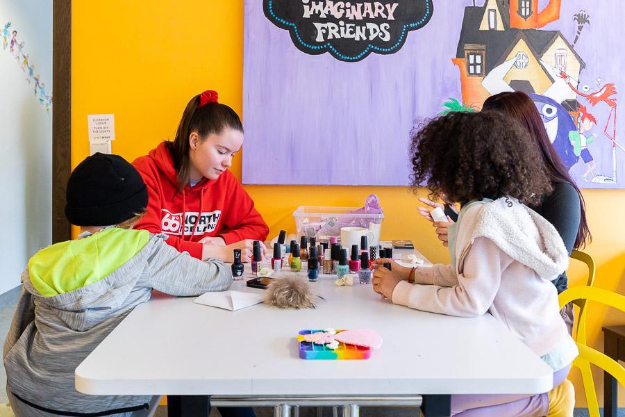 Children sit at a table with an instructor, painting their nails. Behind them is an orange wall and large picture with the heading "Imaginary friends." 