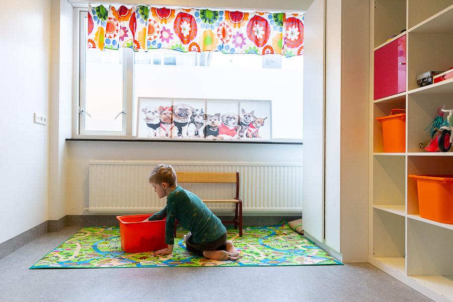 A child sits on a patterned rug playing. Window with colorful curtains in the background. Shelves to the side. 
