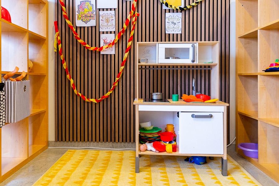 A toy kitchen on a yellow rug. A brown wall with concertina-folded paper decorations in the background. Shelves on both sides. 