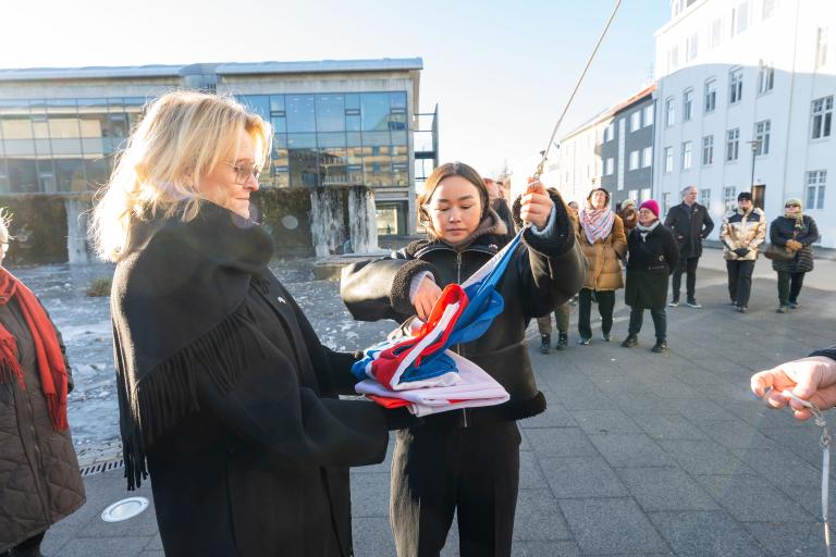 Mayor Heiða Björg Hilmisdóttir and Malu Hendriksen, a Greenlandic intern at Greenland's representative office in Iceland. They hold the Icelandic flag, ready to raise it. Shortly after, the Greenlandic one was hoisted. People in the background, blue sky, winter.