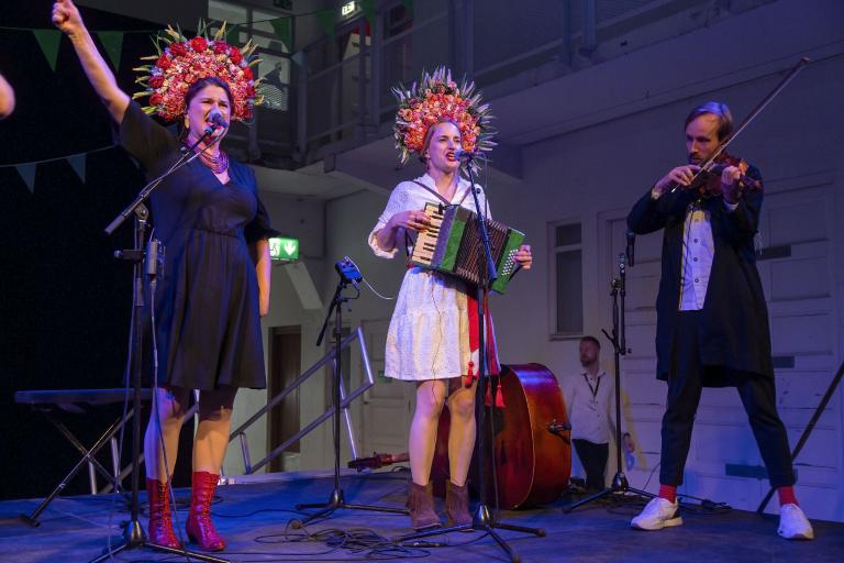 A scene from the opening of the 2024 Reykjavík Arts Festival. Three performers play music on stage—a vocalist, a violinist, and an accordionist in the center. They wear elaborate headdresses.