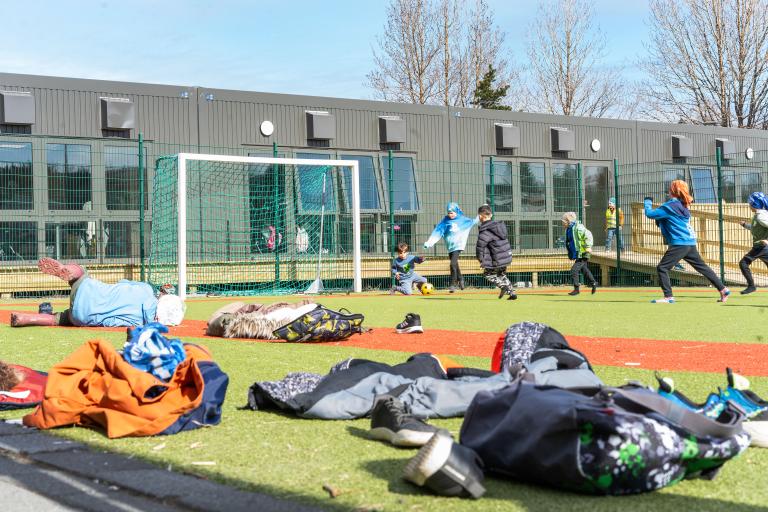 Children play soccer during recess at Ölduselsskóli. Jackets lie on the ground in the foreground while children play soccer in the background. The weather is pleasant.